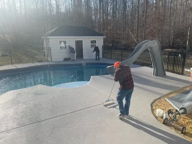 Person skims a backyard pool beside a small pool house and wooded fence line.