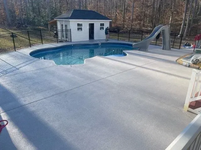 Backyard pool with concrete deck, small shed, and slide beside wooded trees