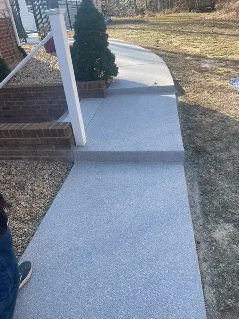 Fresh concrete sidewalk ramp beside a brick porch with white posts and a small evergreen tree