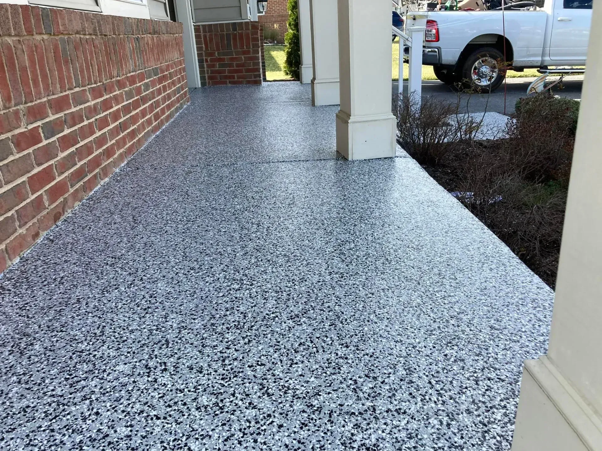 Speckled concrete porch beside a brick wall and beige columns, leading to a parking lot with a white truck.