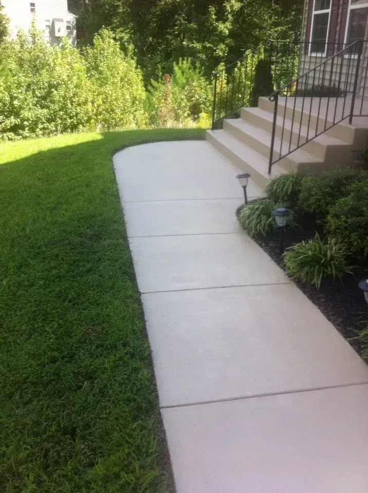 Concrete walkway curving through a green yard beside house steps and garden beds