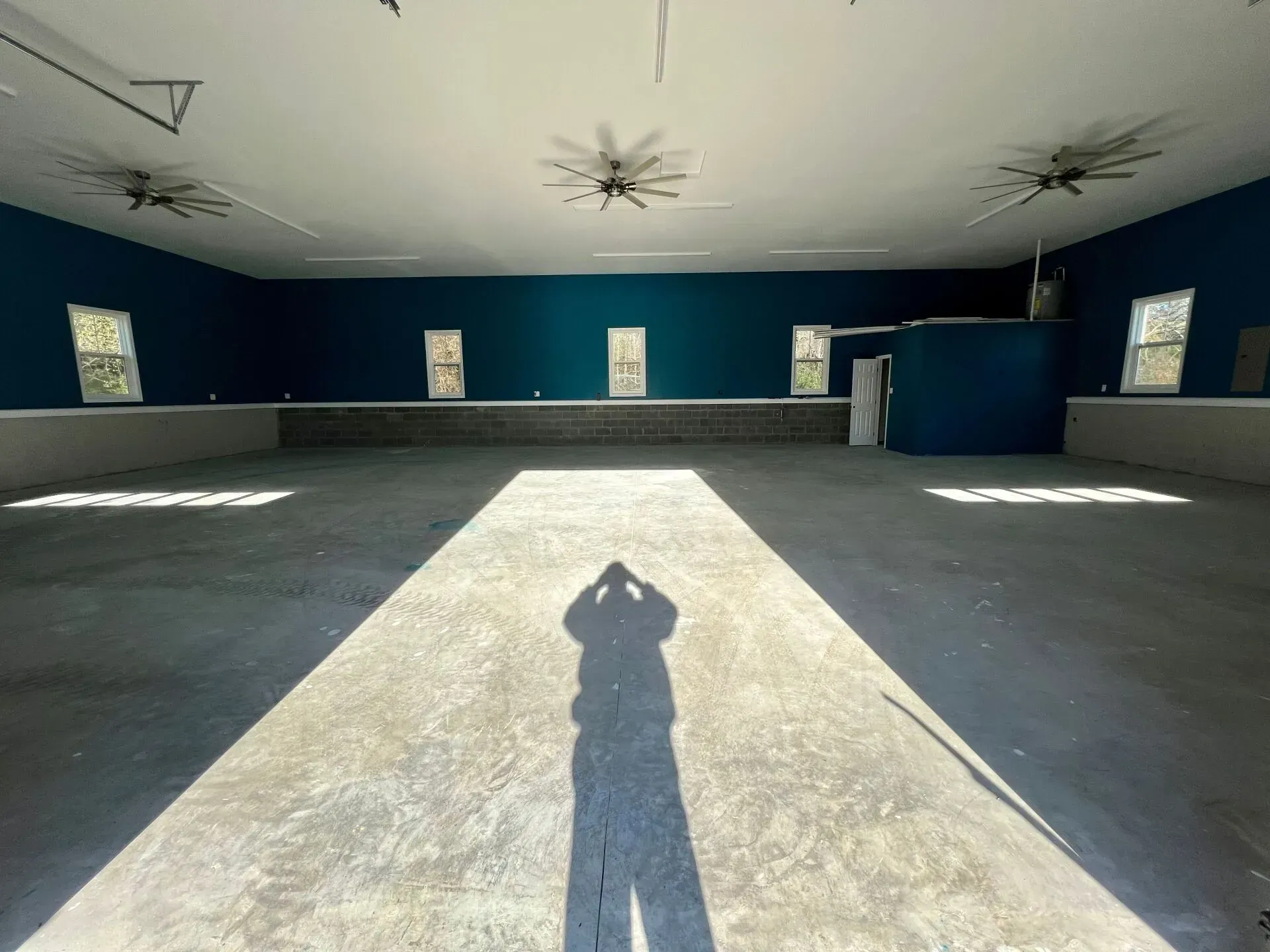 Empty indoor hall with blue walls, high ceiling fans, and sunlight streaming onto a central walkway.