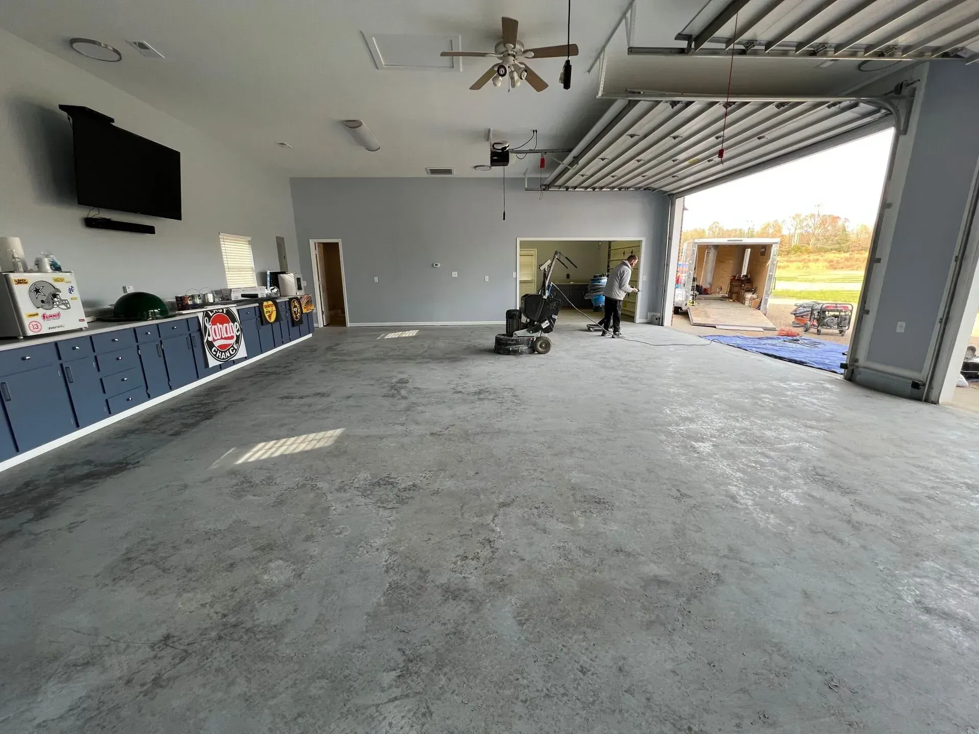 Spacious garage with gray floor, wall-mounted TV, workbench, and open overhead door to a sunny driveway.