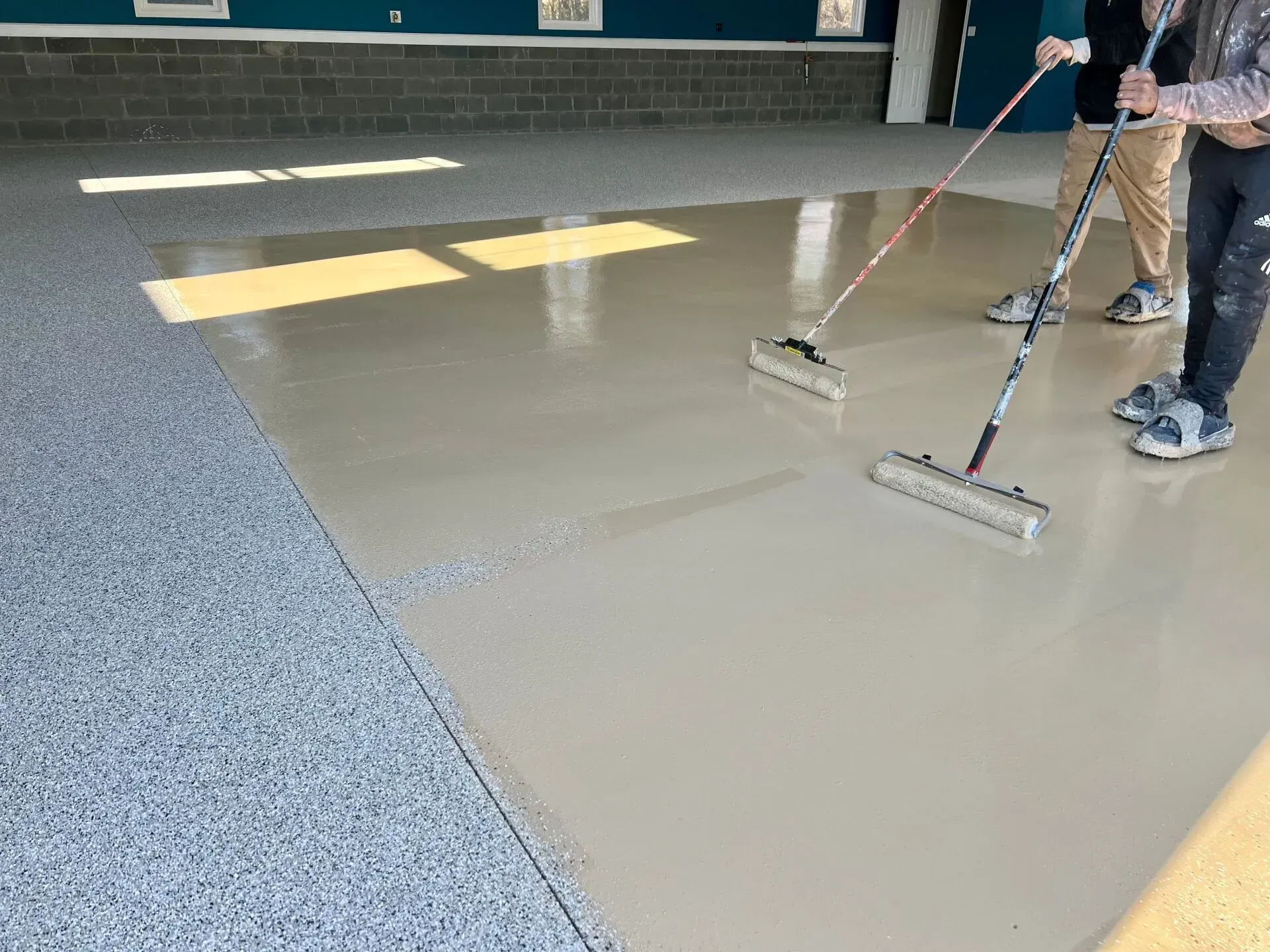 Two workers smoothing wet gray flooring with squeegees in a building hallway