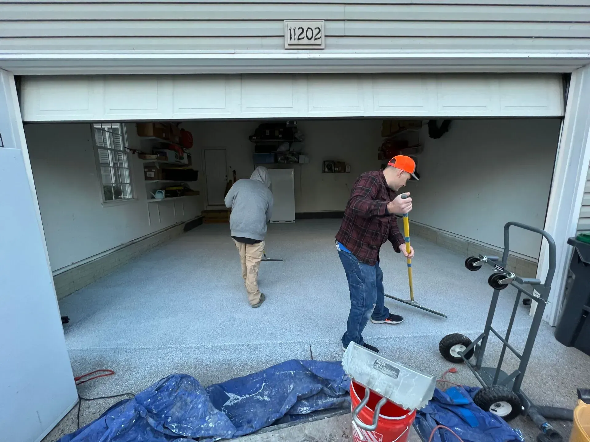 People cleaning a garage floor, one mopping and one sweeping, with tools and supplies scattered around.