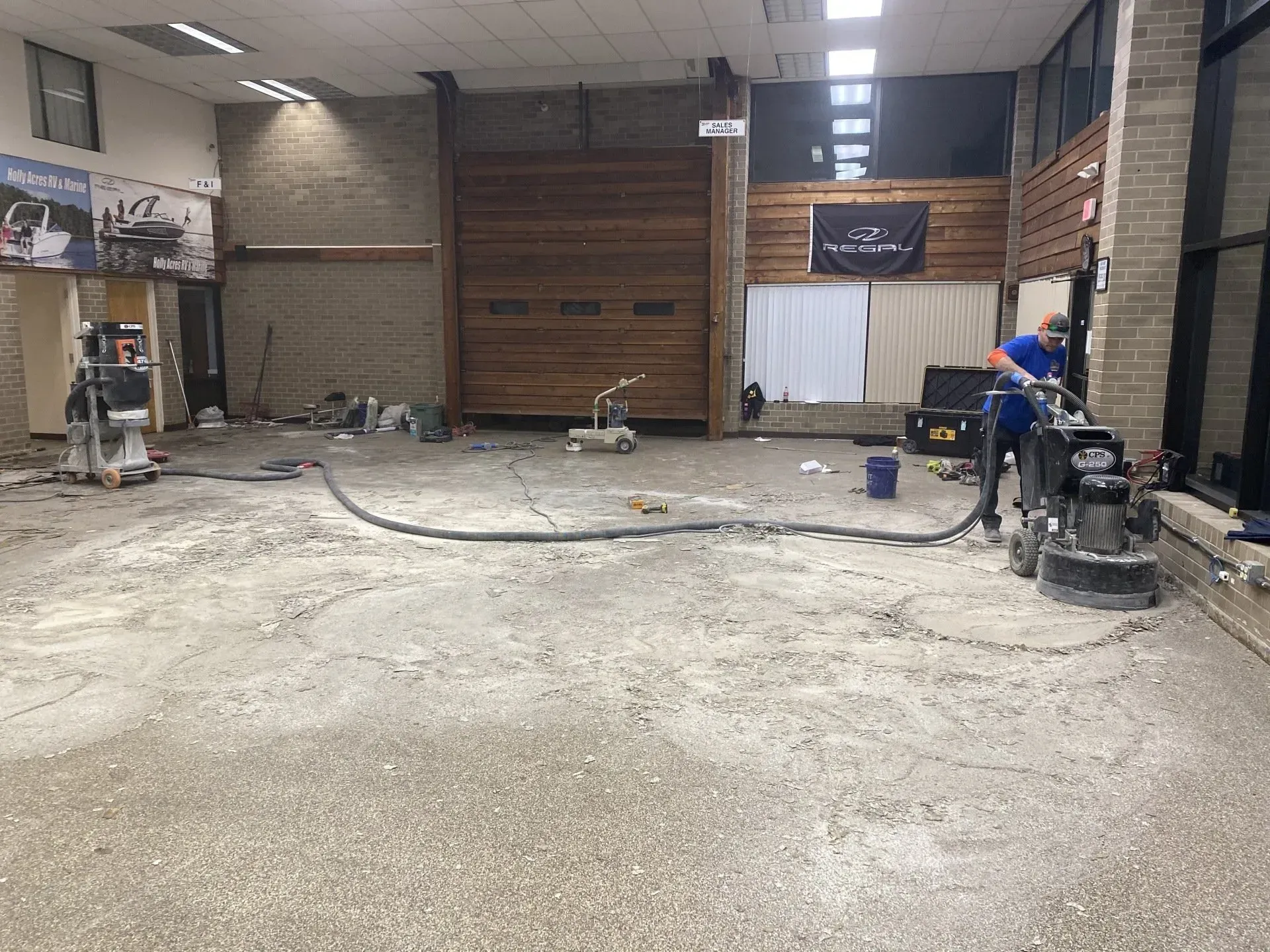 Large empty hall with unfinished concrete floor, tools, and a worker on the right near the wall.
