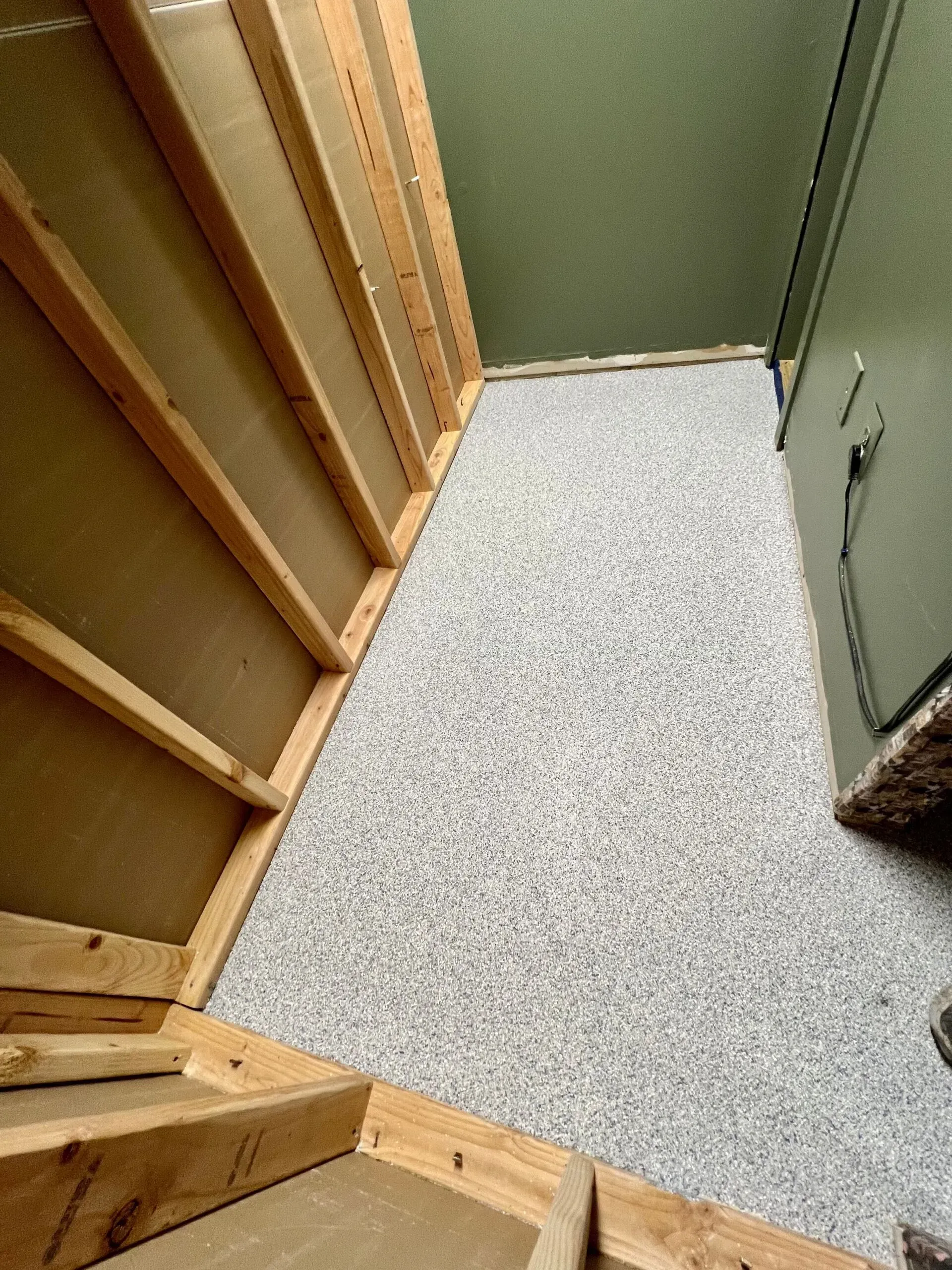 Narrow unfinished room with wood framing, speckled floor, and olive-green walls.