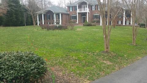Suburban brick house with a large front lawn, trees, and a driveway on an overcast day