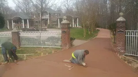 Workers repairing a red driveway beside a gated entrance and brick pillars, with a house in the background