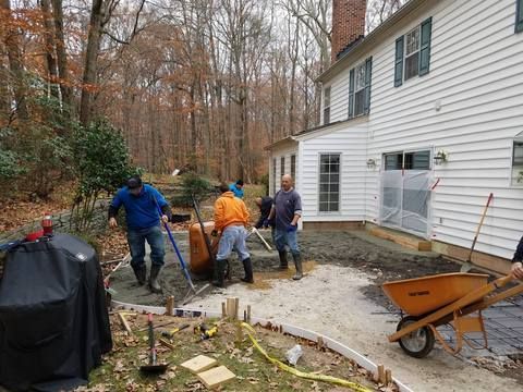 Workers paving a backyard patio beside a white house, with tools, wheelbarrow, and covered grill.