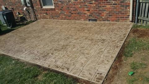Concrete patio slab beside a brick house with a stamped stone pattern and grass around the edges