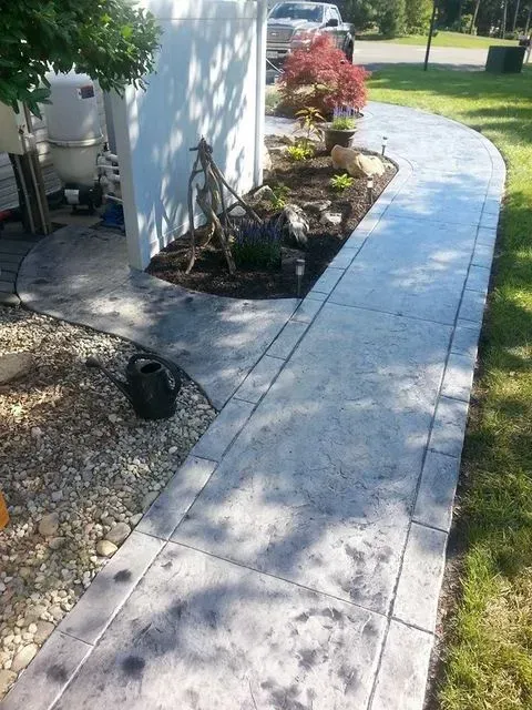 Curved concrete walkway beside a house, bordered by gravel, grass, and small garden plants.