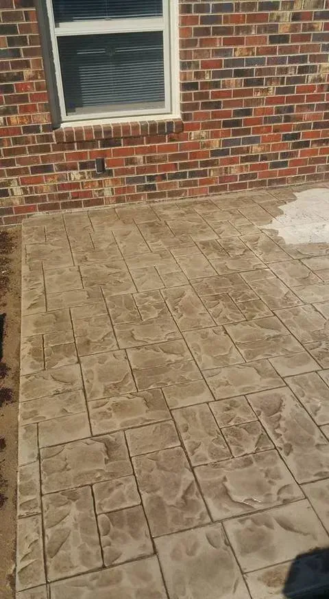 Stamped concrete patio beside a brick house wall and window, with a small white patch on the right.