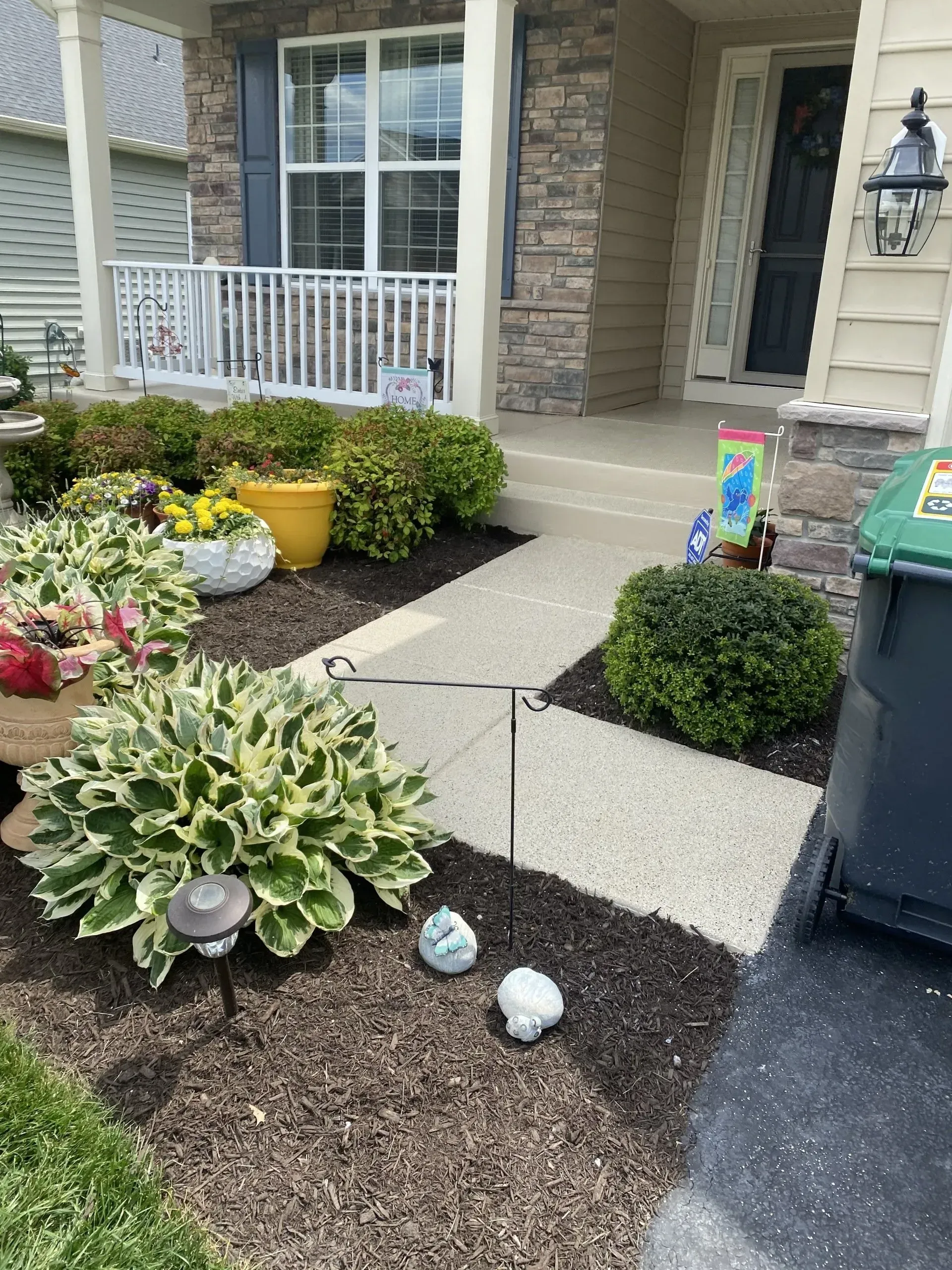 Front walkway with potted flowers, hostas, and trimmed shrubs beside a house porch