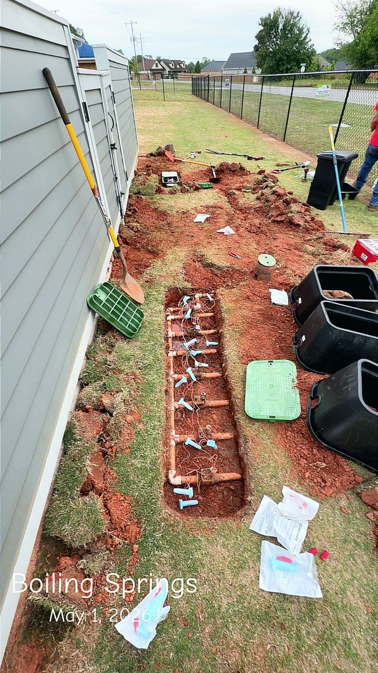 Backyard garden bed alongside a house, with red soil, brick edging, and scattered tools and bins