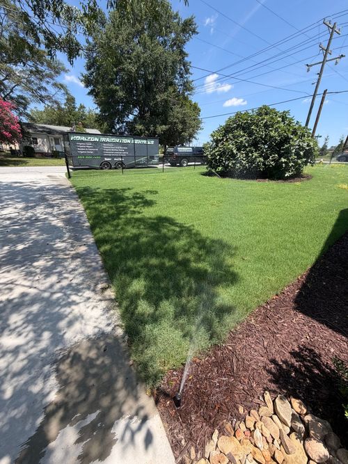 Sunny lawn and sidewalk in a landscaped park with trees, bushes, and a sign in the background