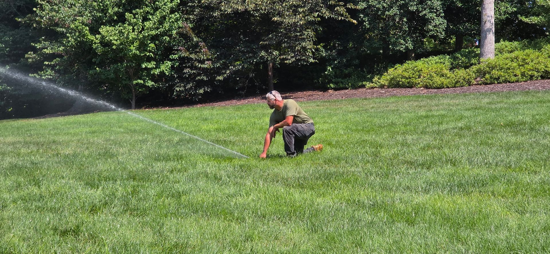Person kneeling on a grassy lawn near a sprinkler spraying water