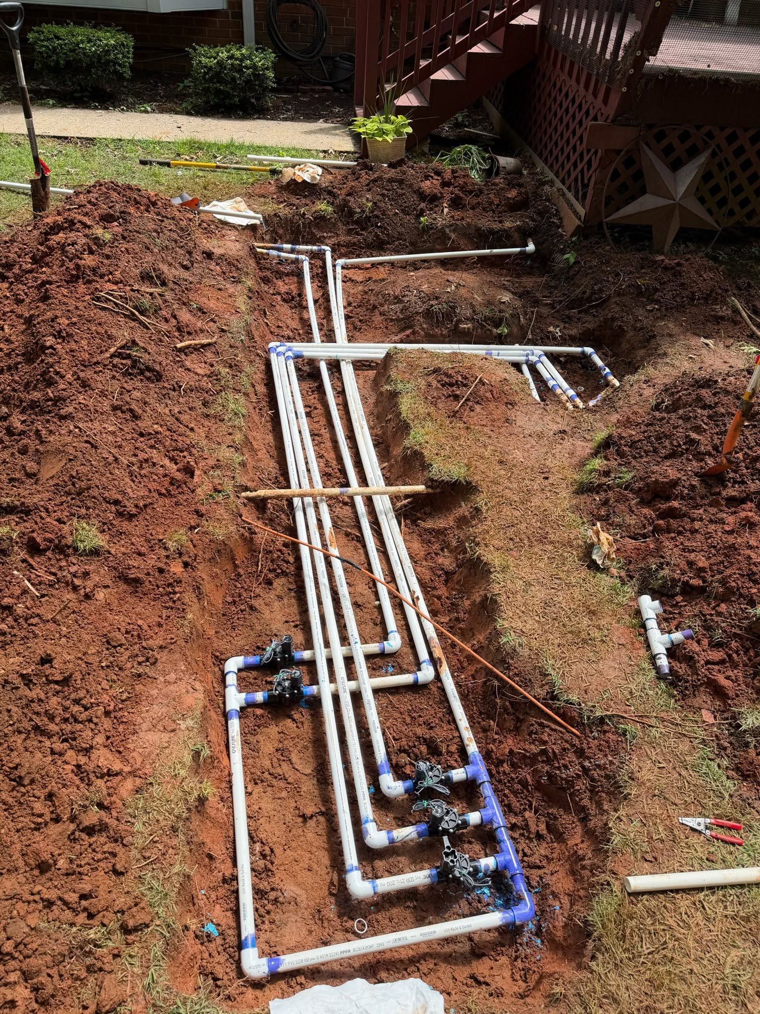 PVC pipes laid in trenches in a dirt yard, forming a plumbing layout with blue fittings.