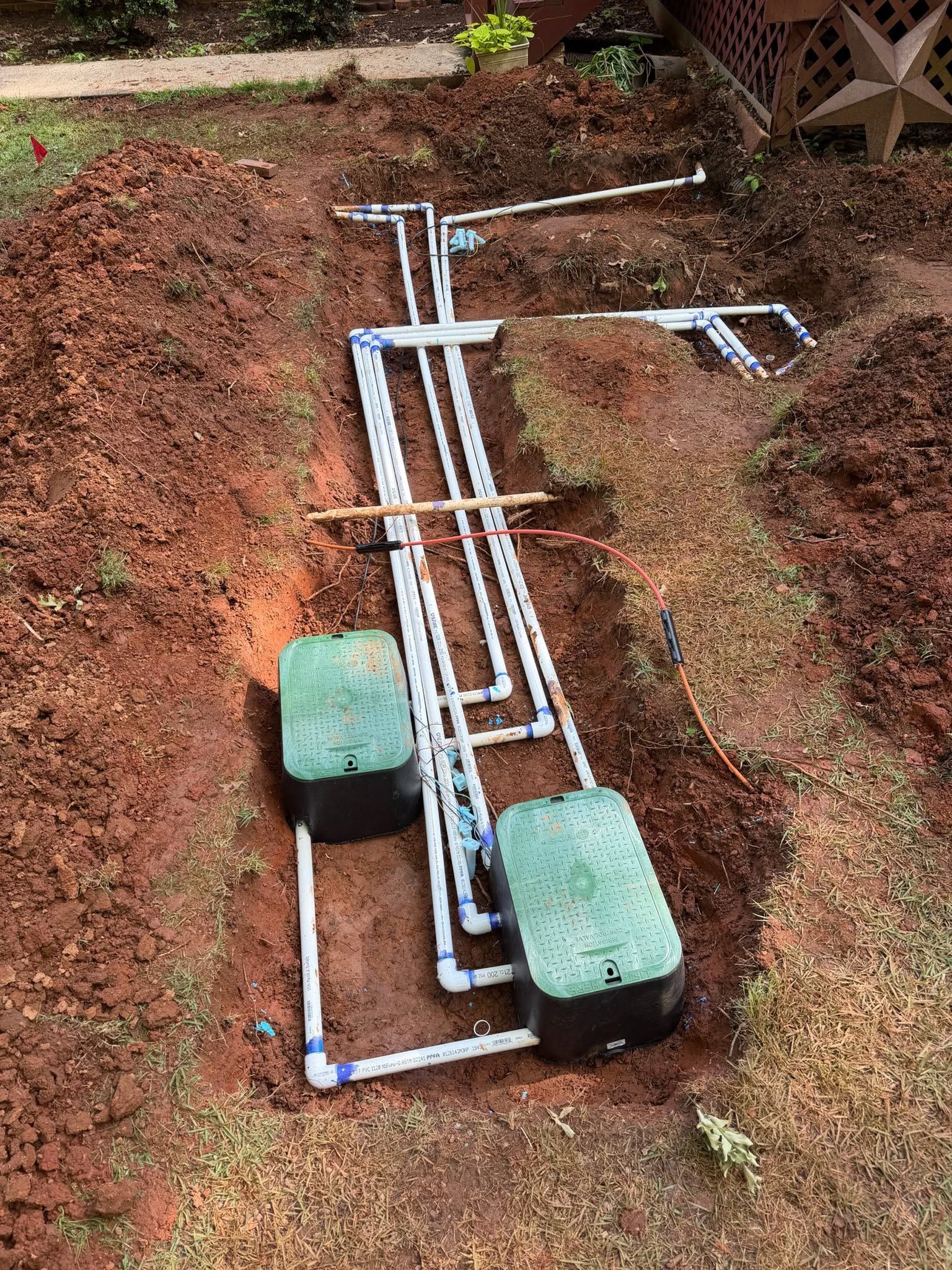 A shallow trench with exposed white PVC pipes beside two green bricks on bare soil.