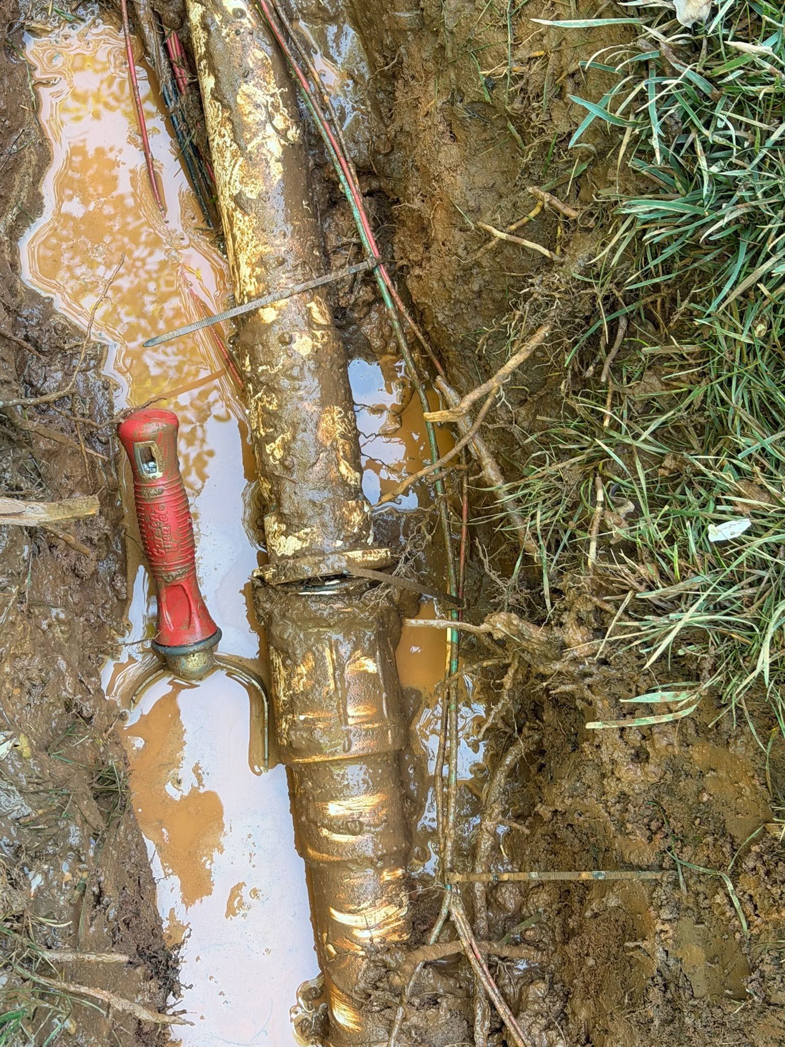 Rusty underground pipes beside muddy soil and grass, with a red valve handle visible.