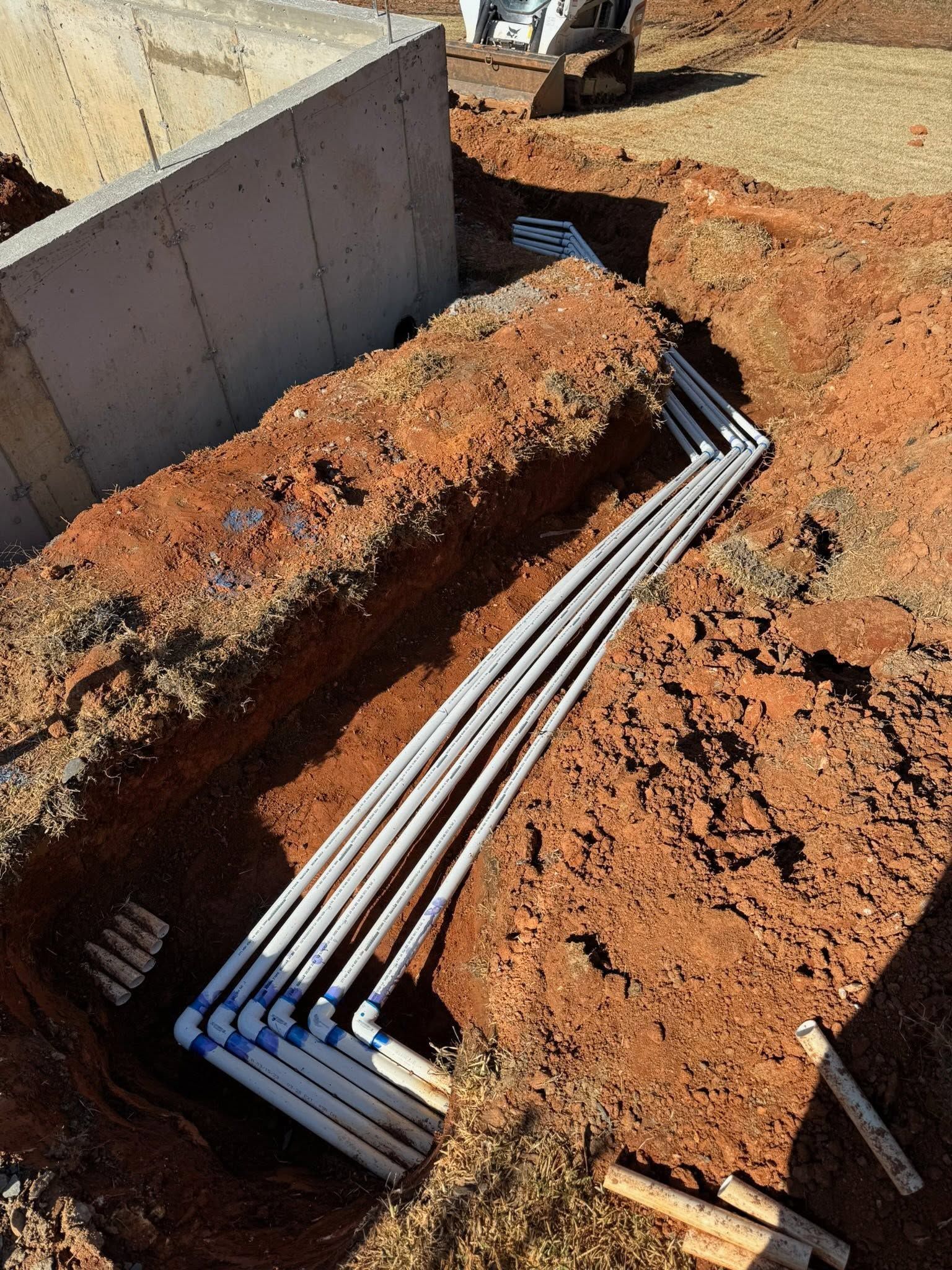 Underground electrical conduits laid in a dirt trench beside a concrete foundation wall.