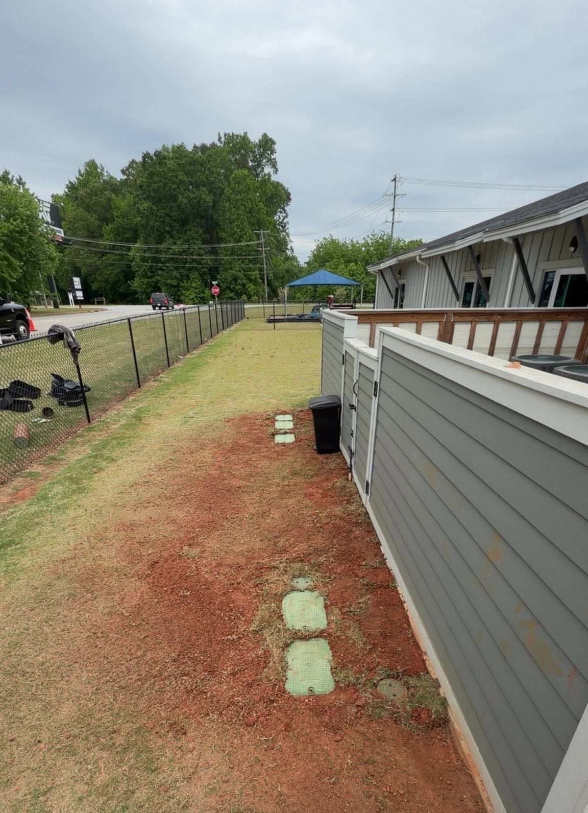 Narrow grassy side yard beside a gray house, with a fence, trees, and a paved road in the distance.