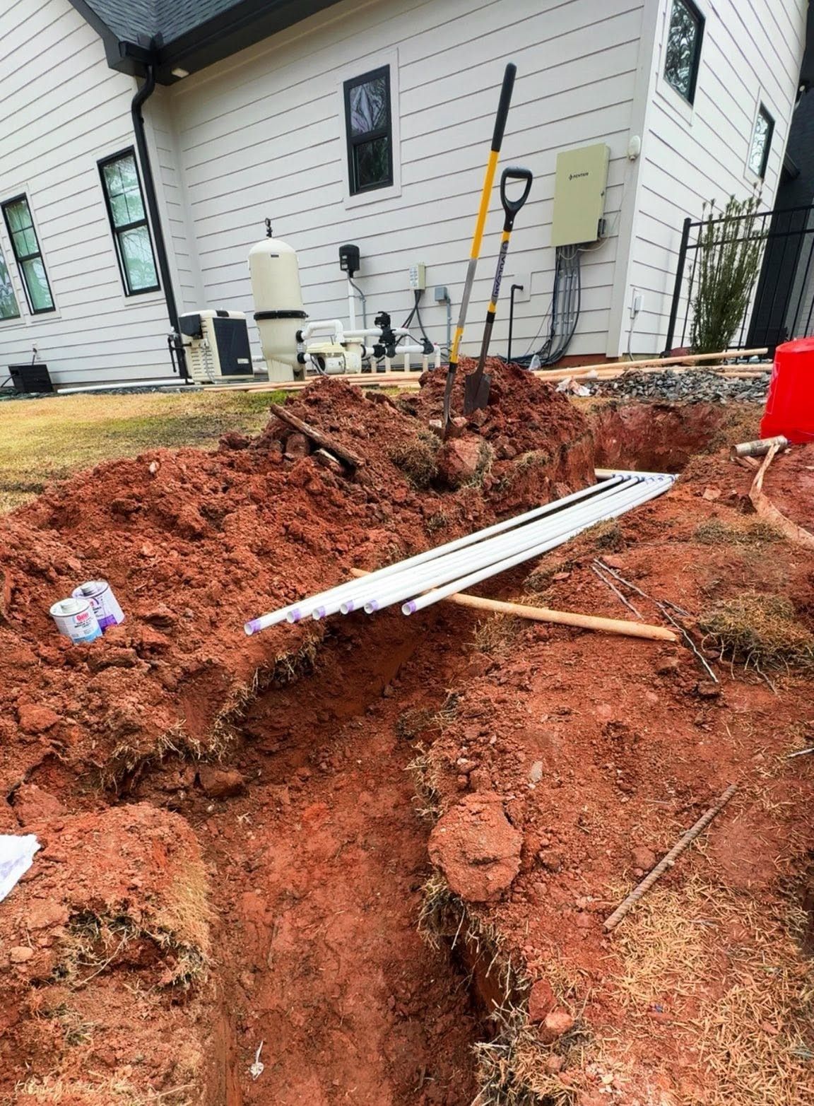 Backyard trench beside a house, with red dirt, a metal sheet, and a shovel in the ground.