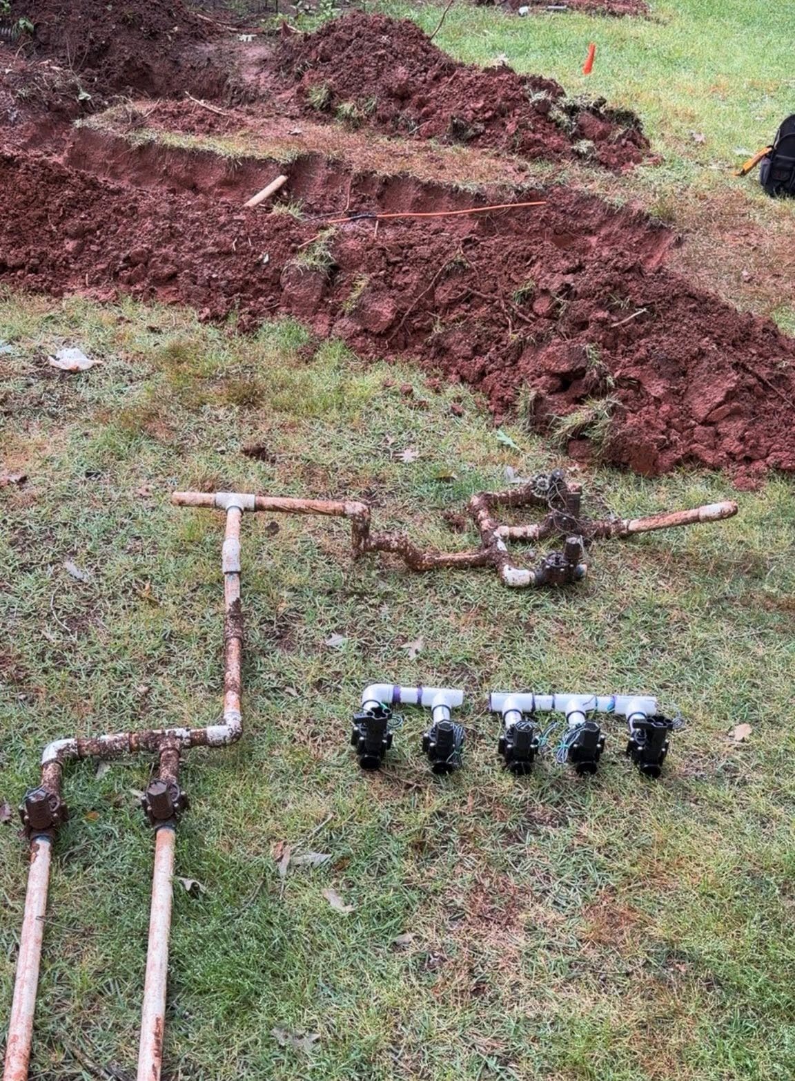 Outdoor trench with exposed pipes and a row of black sprinkler heads on grass.