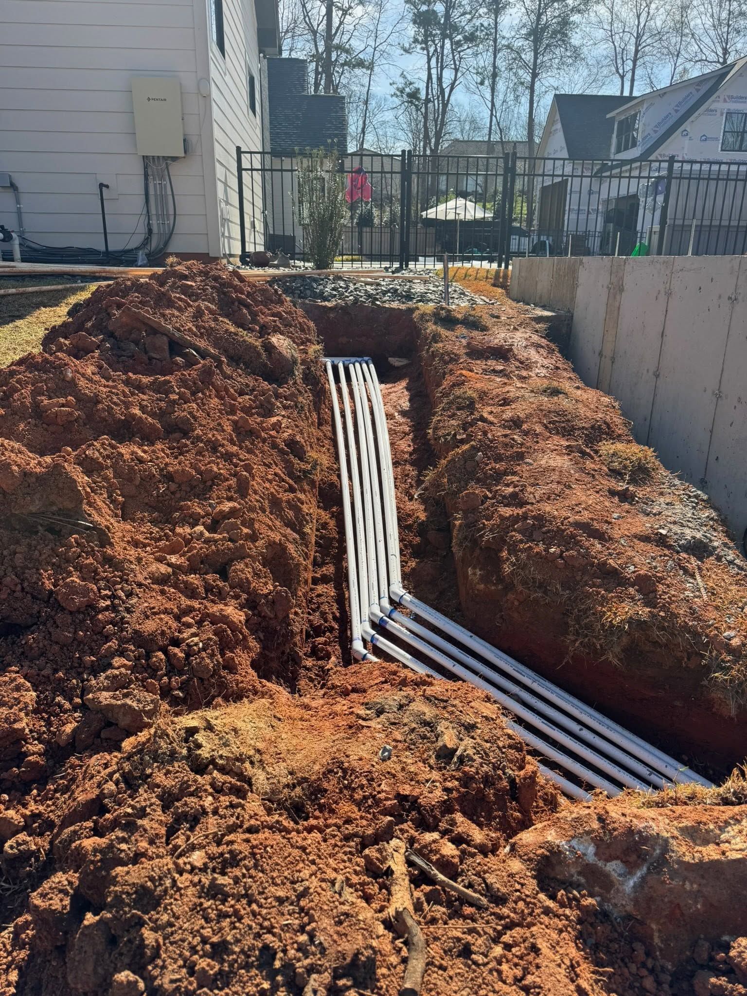 Trenches with bundled white cables laid in red dirt at a construction site beside a fence and houses