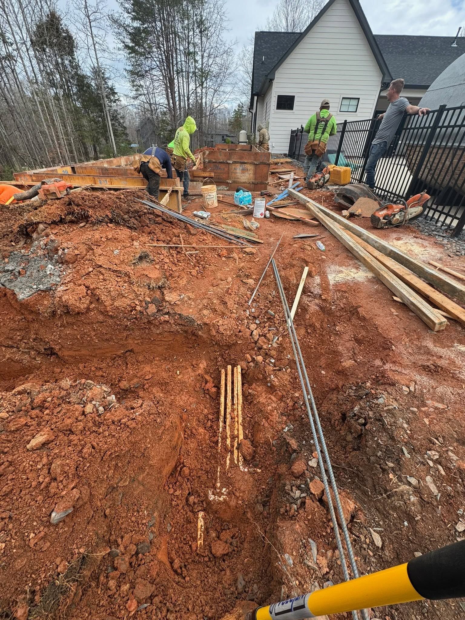 Construction workers on a muddy residential site with excavated soil, pipes, and a house in the background