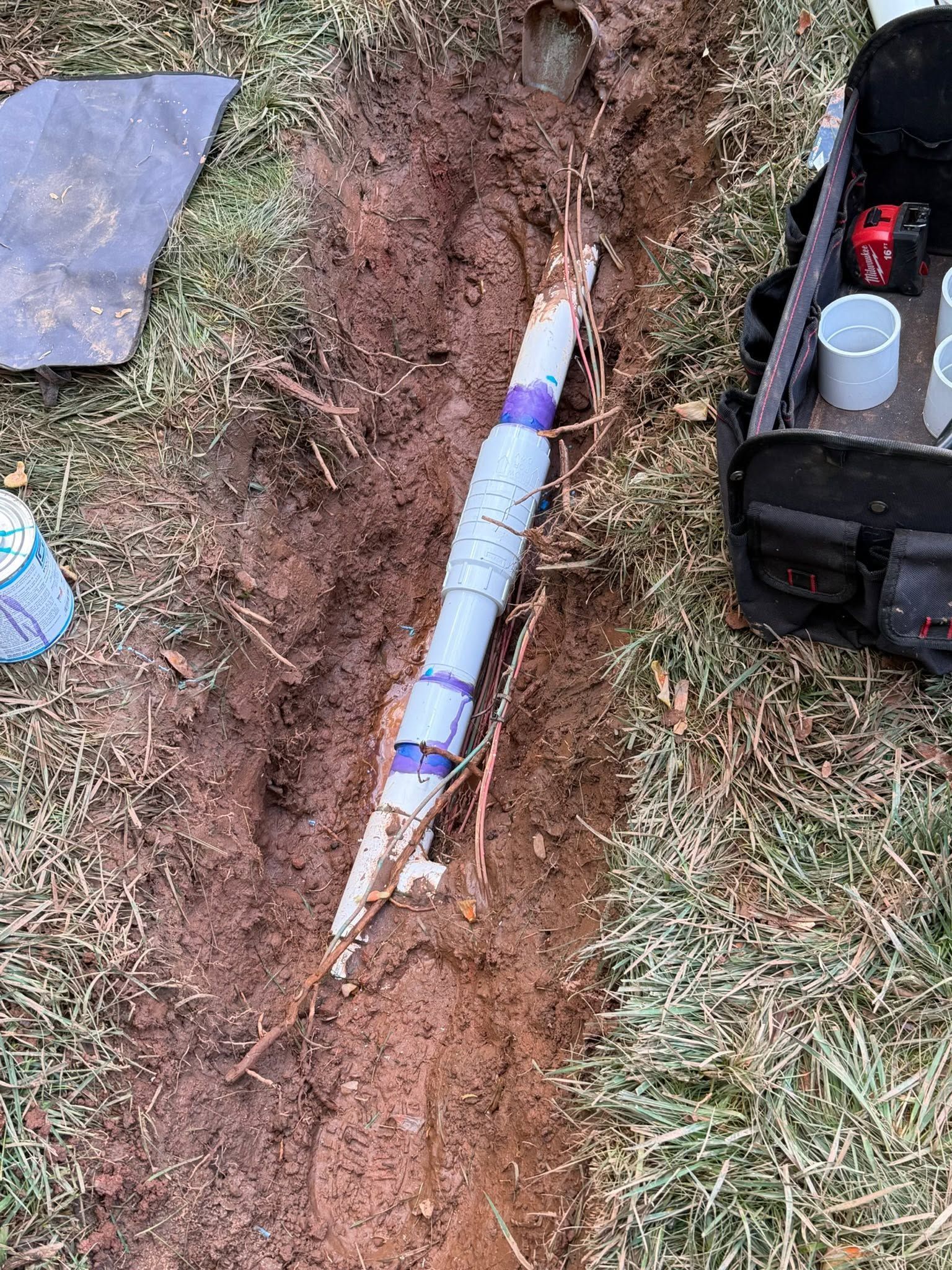 Blue-and-white cylindrical object buried diagonally in a dirt trench beside grass and tools