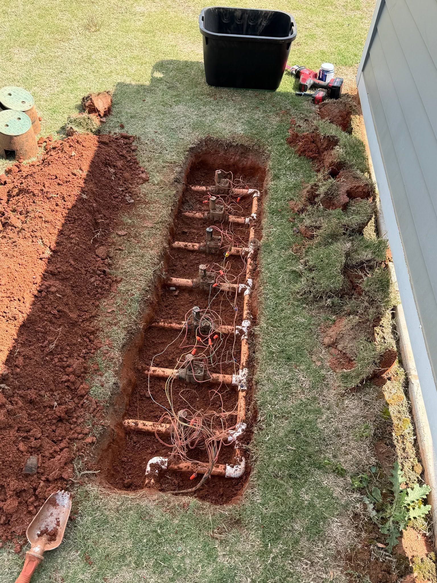 Backyard trench with exposed soil, bordered by grass and mulch, beside a house and a black planter.