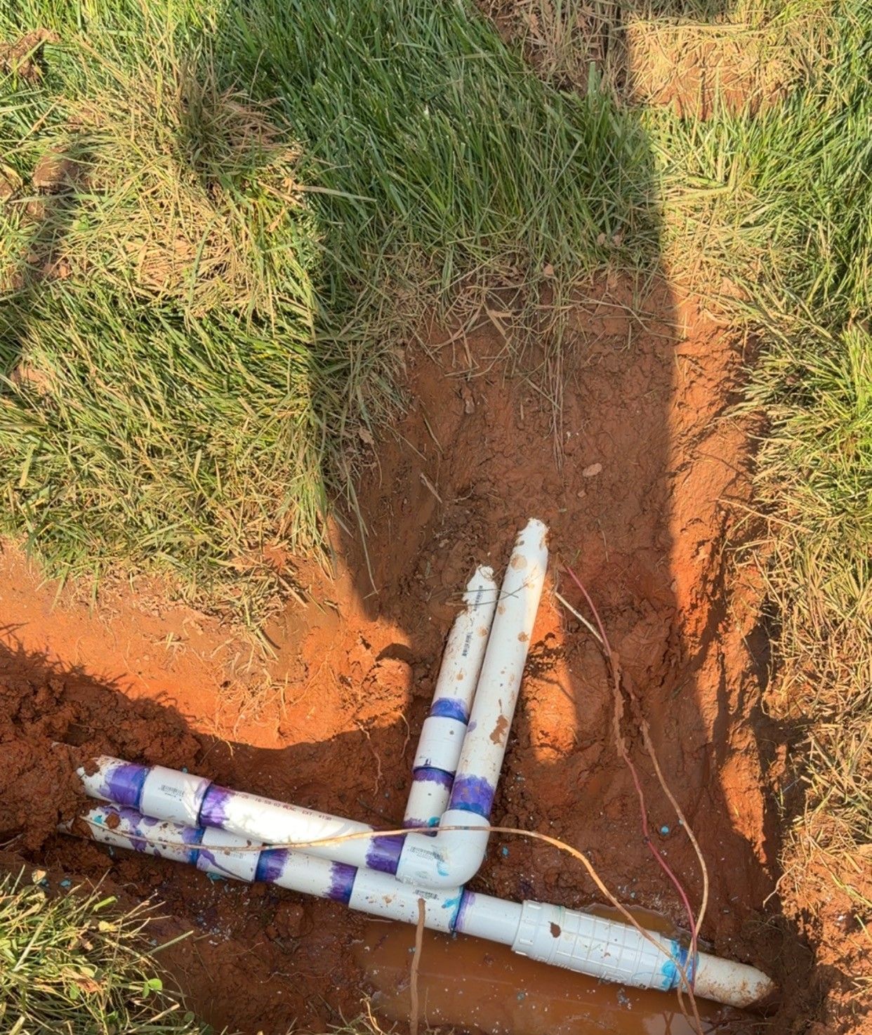PVC pipes exposed in a red dirt trench beside grass, with taped joints and a cable nearby