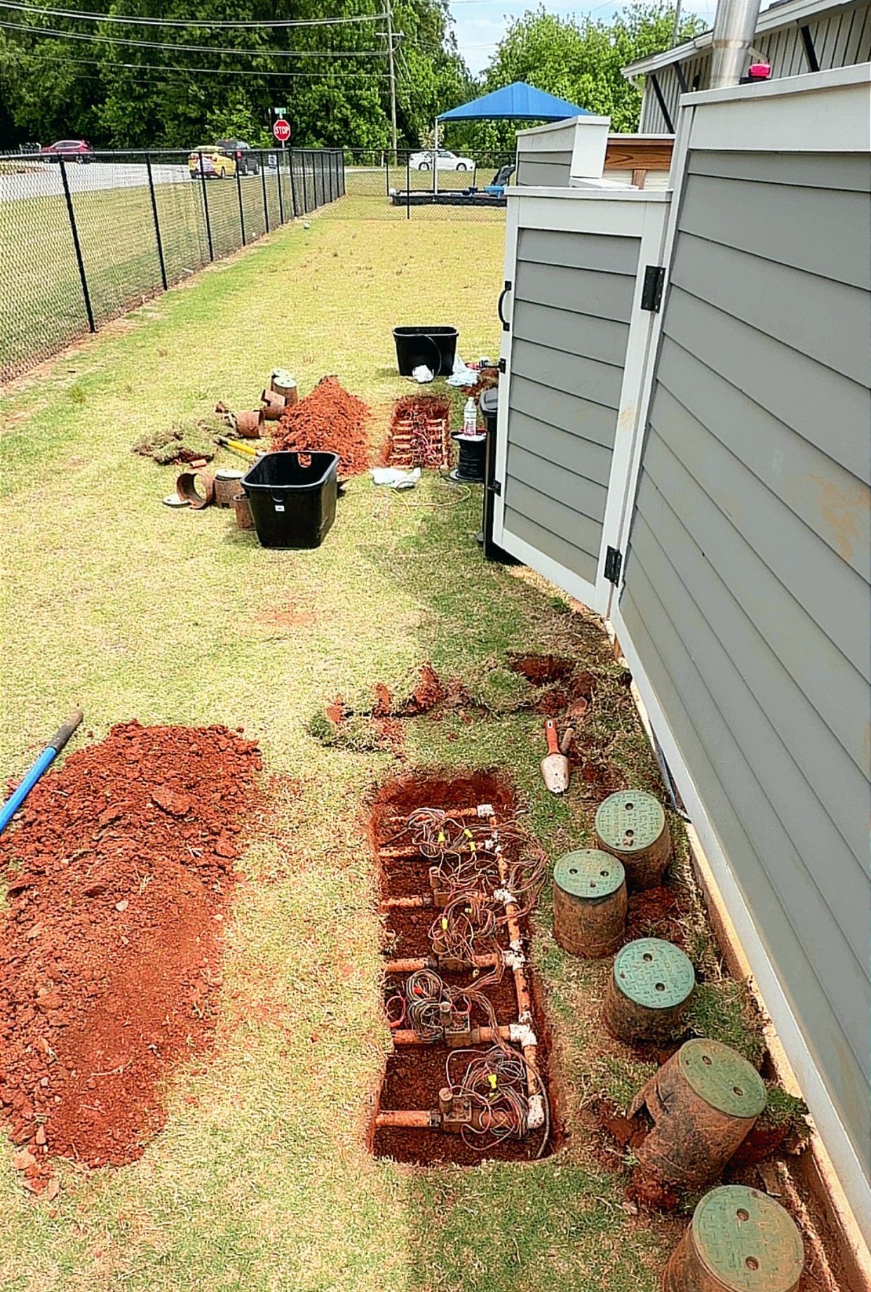 Side yard garden bed with red mulch, potted plants, and a house wall along a fence line.