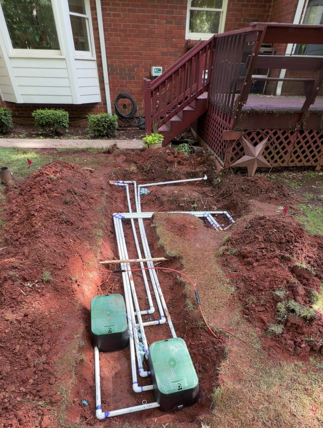 Trenches with white PVC pipes and two green covers in a yard beside a brick house and wooden deck