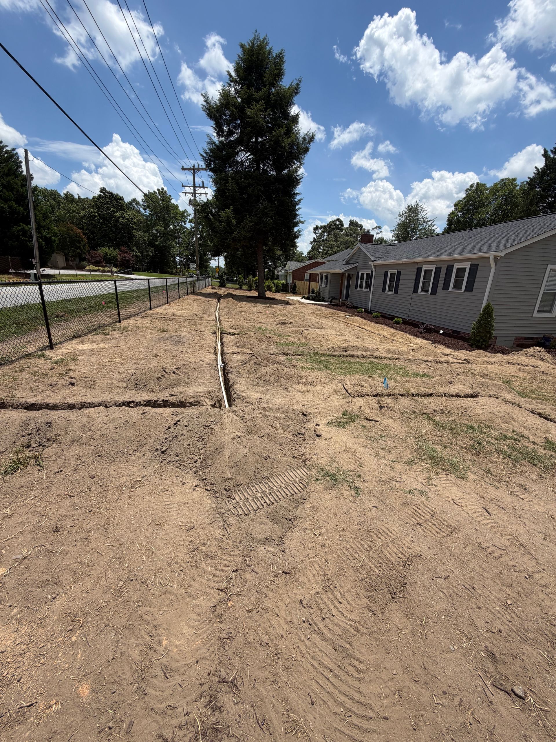 Recently graded dirt lot beside a fence and house under a partly cloudy sky