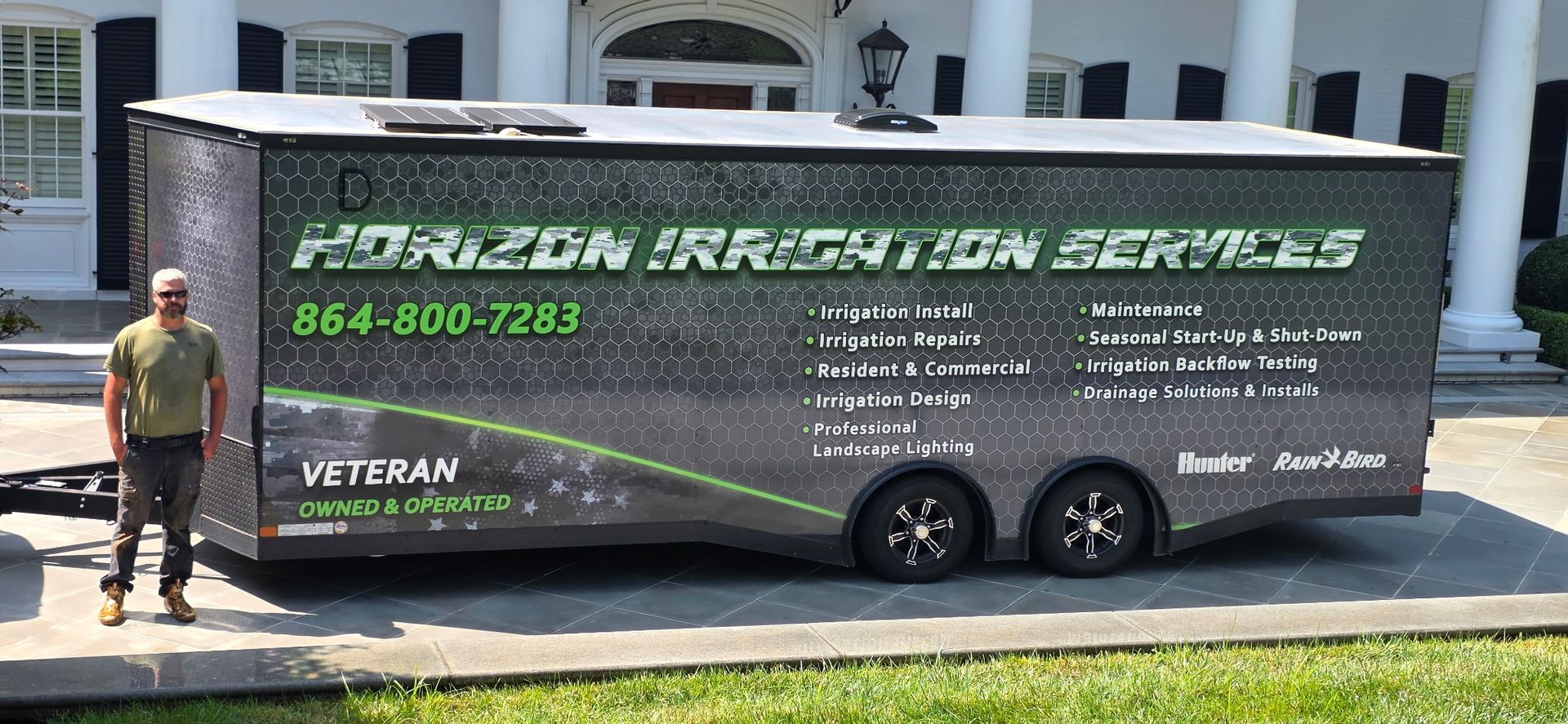 Mobile trailer for lawn care services parked by a house, with a worker standing beside it.