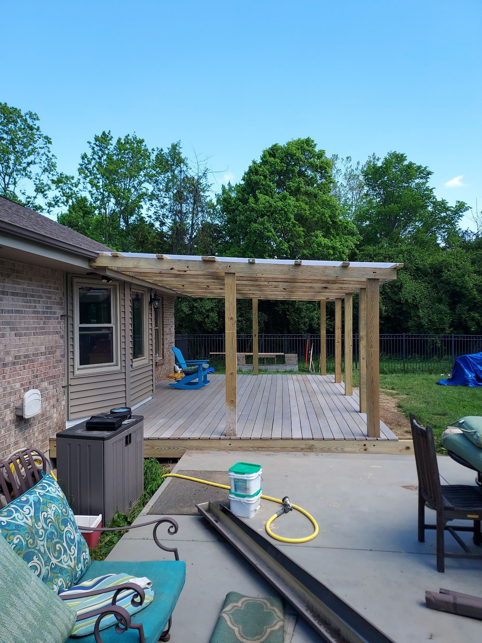 Back patio under construction with wooden pergola, chairs, grill, and tools beside a brick house