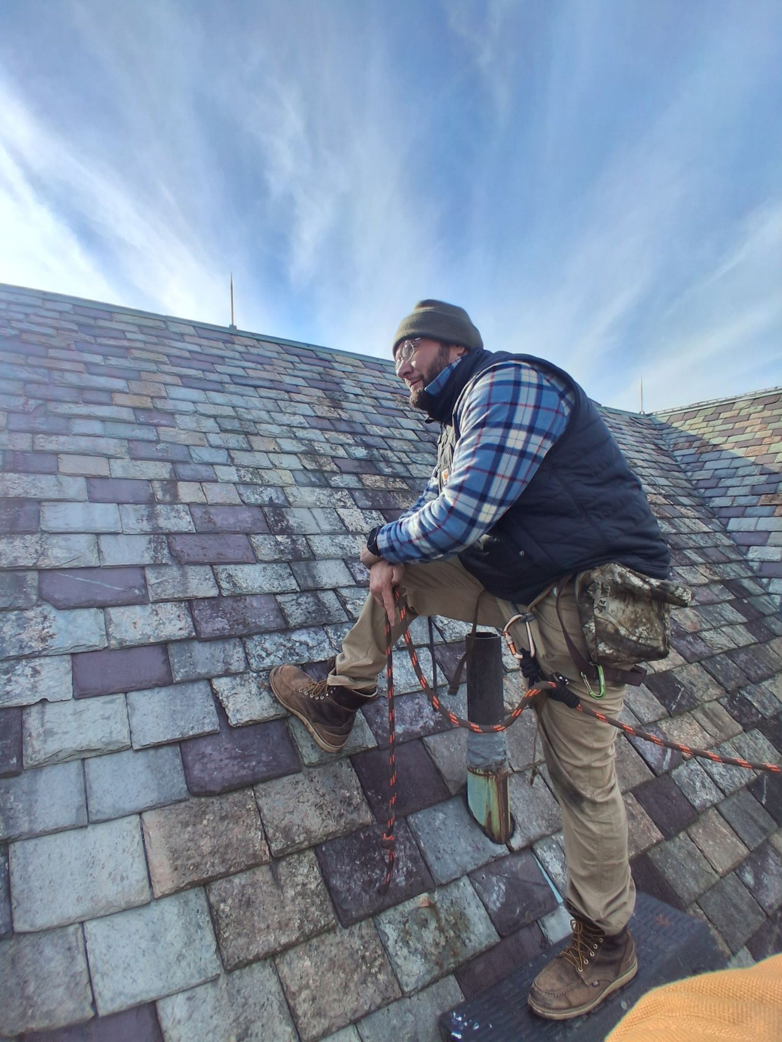 Worker on a steep shingled roof, wearing safety gear and using a harness.