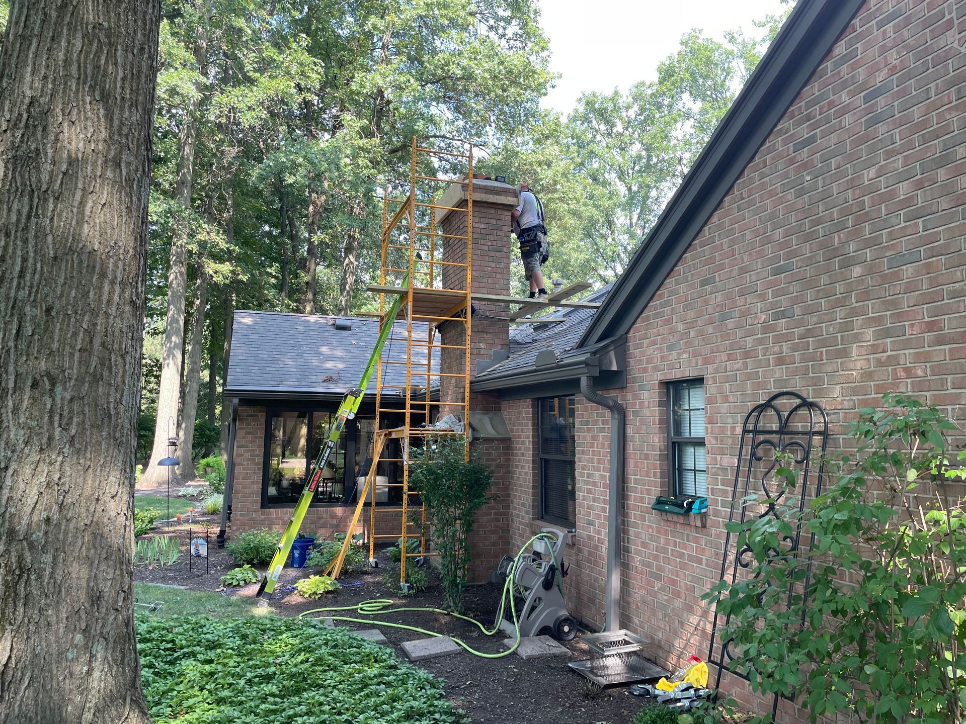 Worker on a roof beside a brick house, using yellow scaffolding and ladder in a wooded yard.