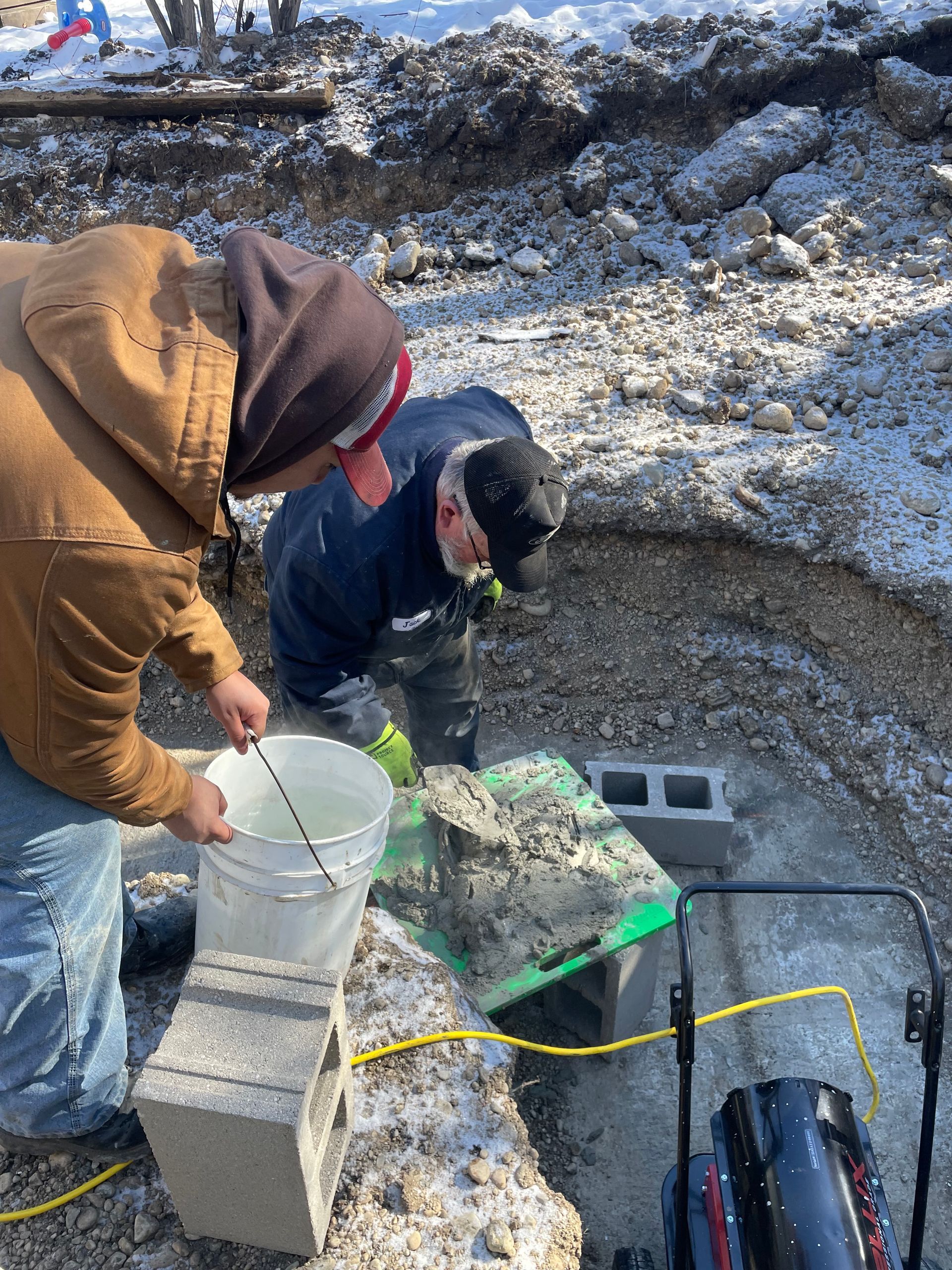 Two workers in a rocky trench handling tools and a bucket beside cinder blocks and cables