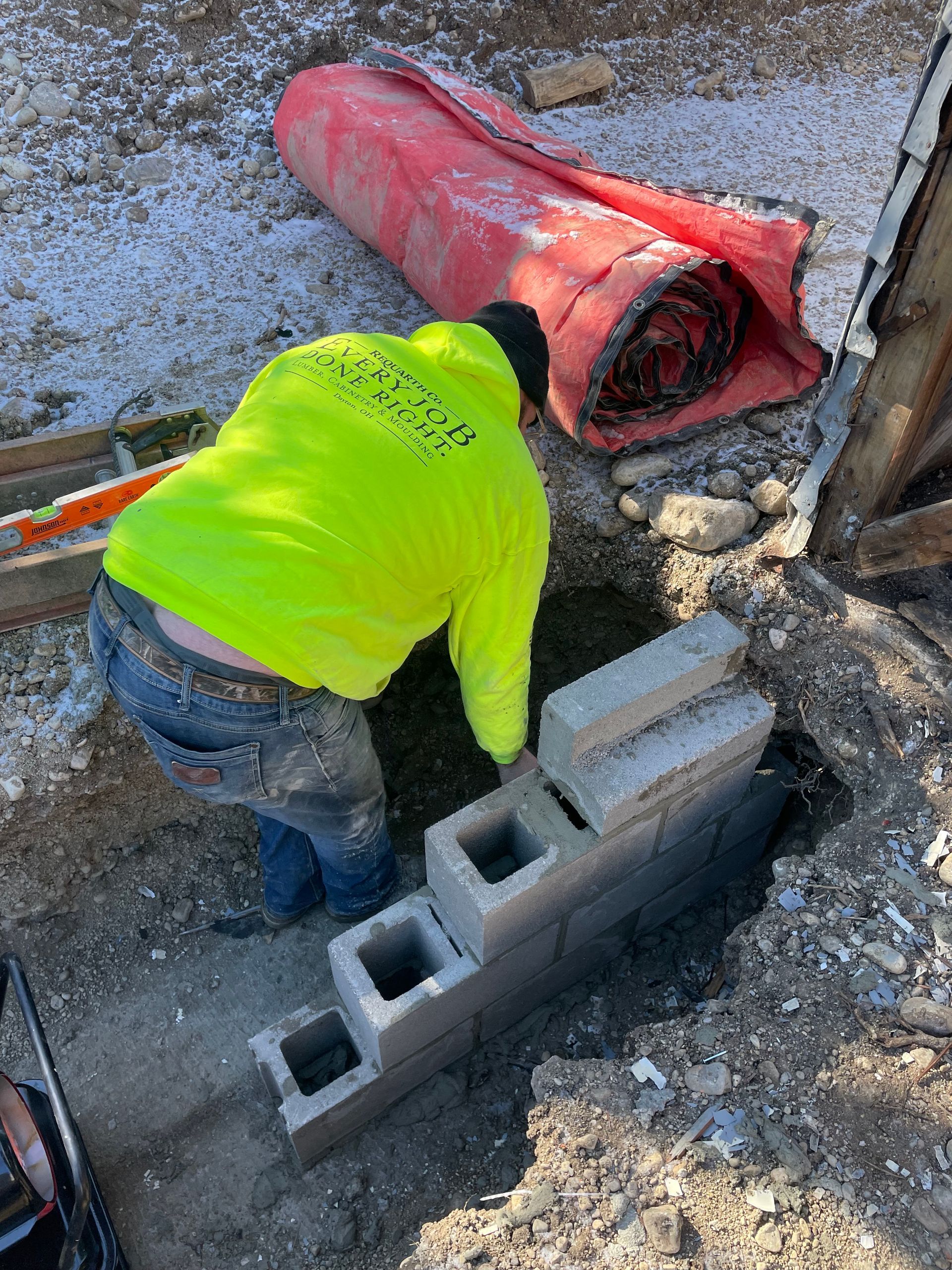 Worker in neon yellow hoodie laying cinder blocks beside a red pipe on a gravel construction site
