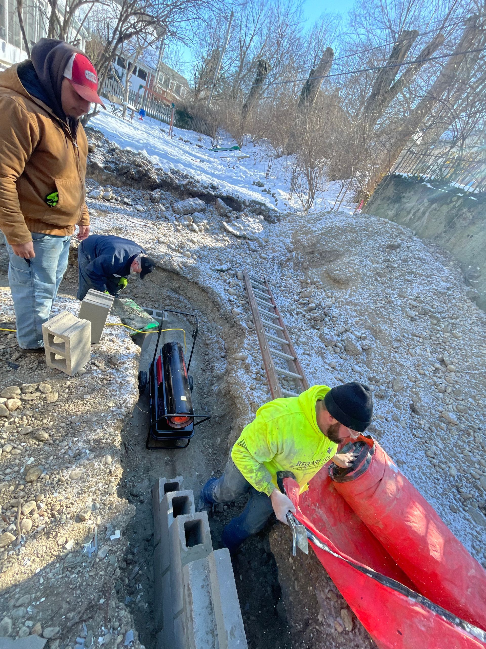 Workers handling a red pipe in a snowy roadside ditch beside cinder blocks and tools