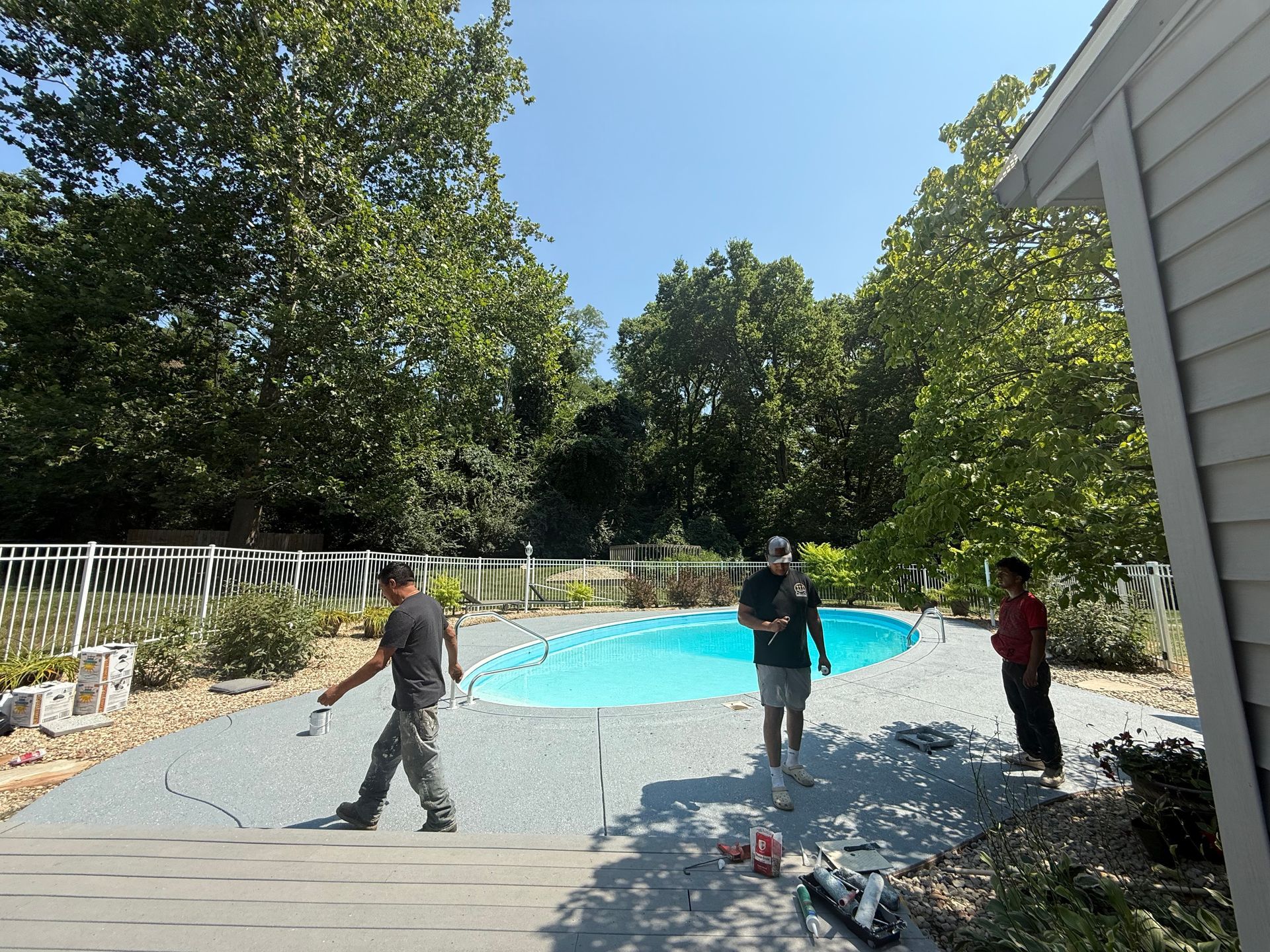 People working around an above-ground pool in a backyard, with a white fence and trees.