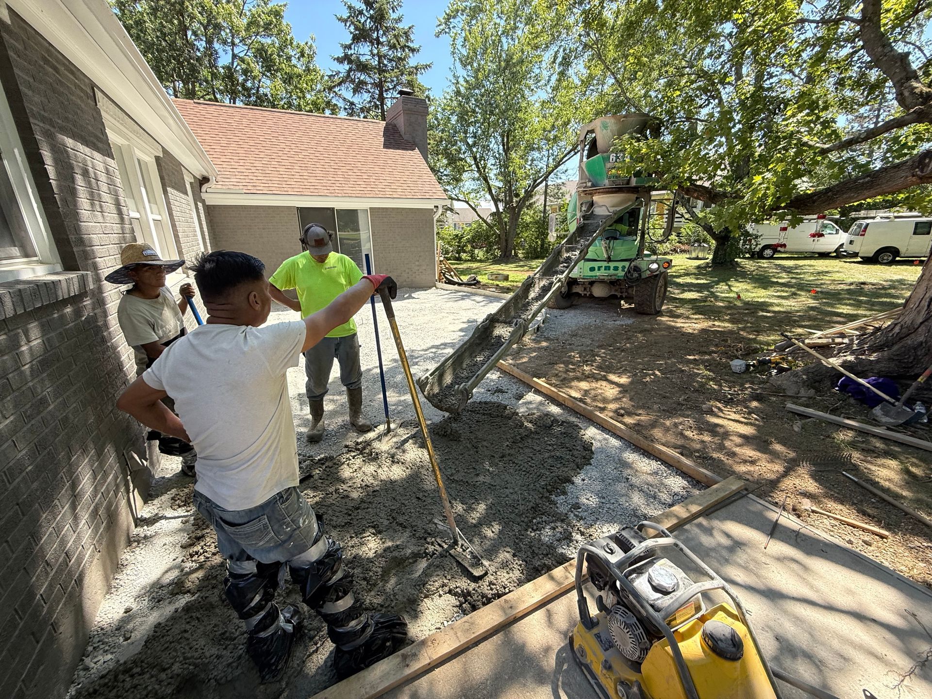 Workers pour concrete beside a house while another operates a machine in the yard.