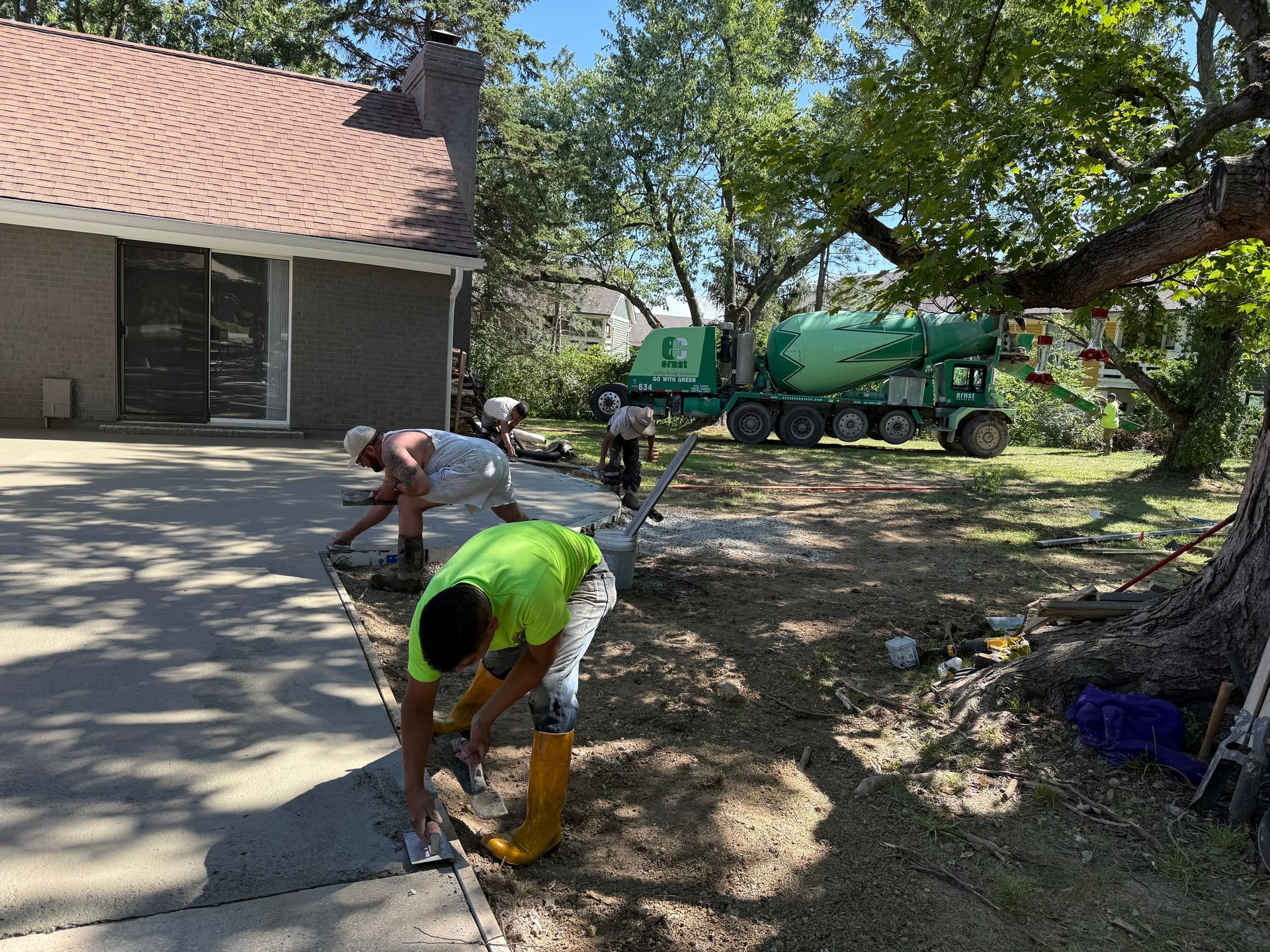 Workers in bright safety gear repair a driveway beside a house, with a green utility truck nearby.