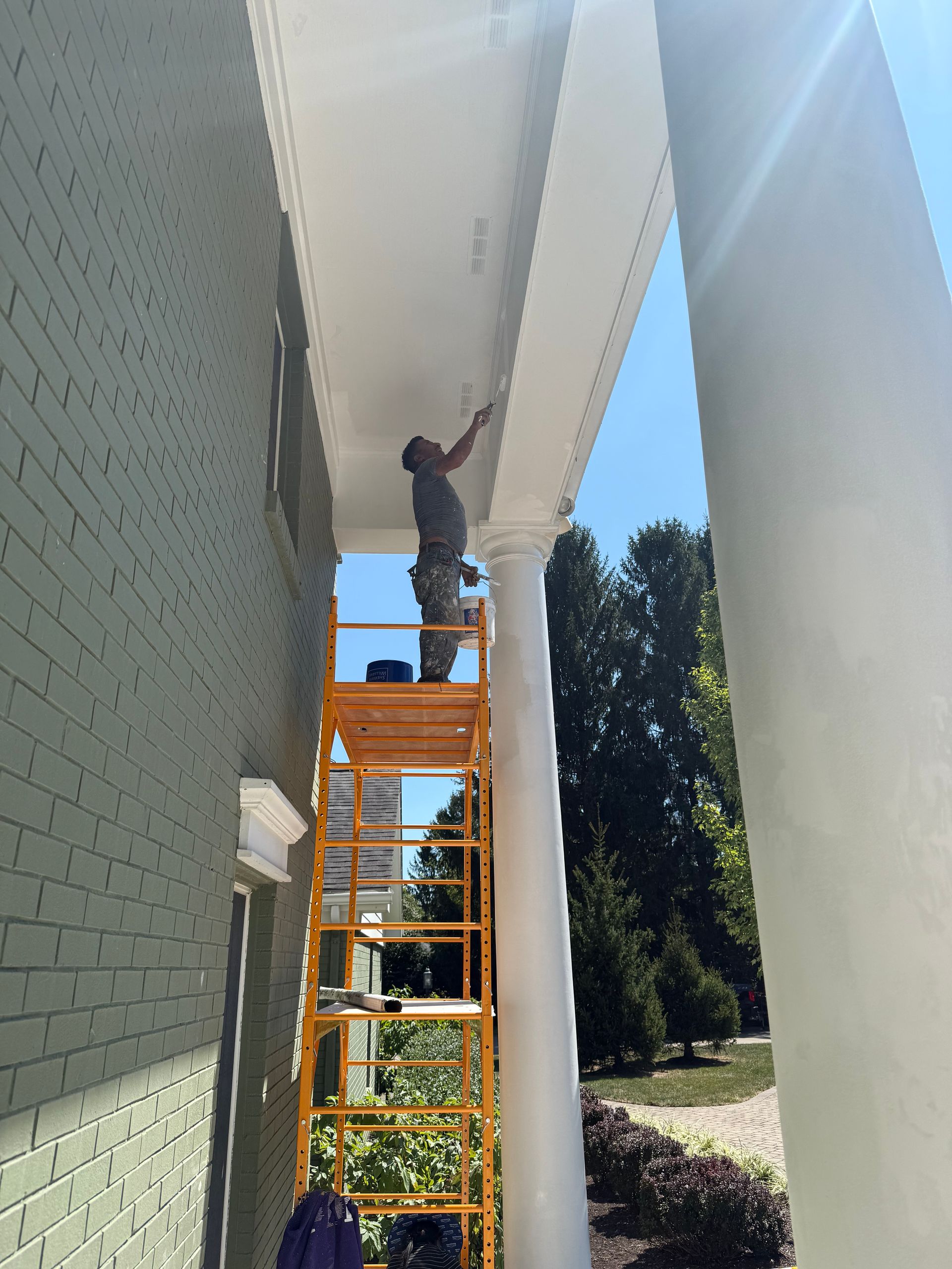 Worker standing on an orange ladder reaching up under a white roof eave beside a brick wall