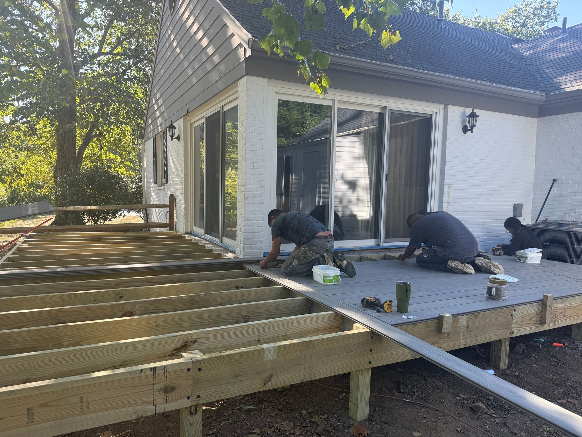 Workers building a wooden deck beside a white house, with tools and materials on the platform.