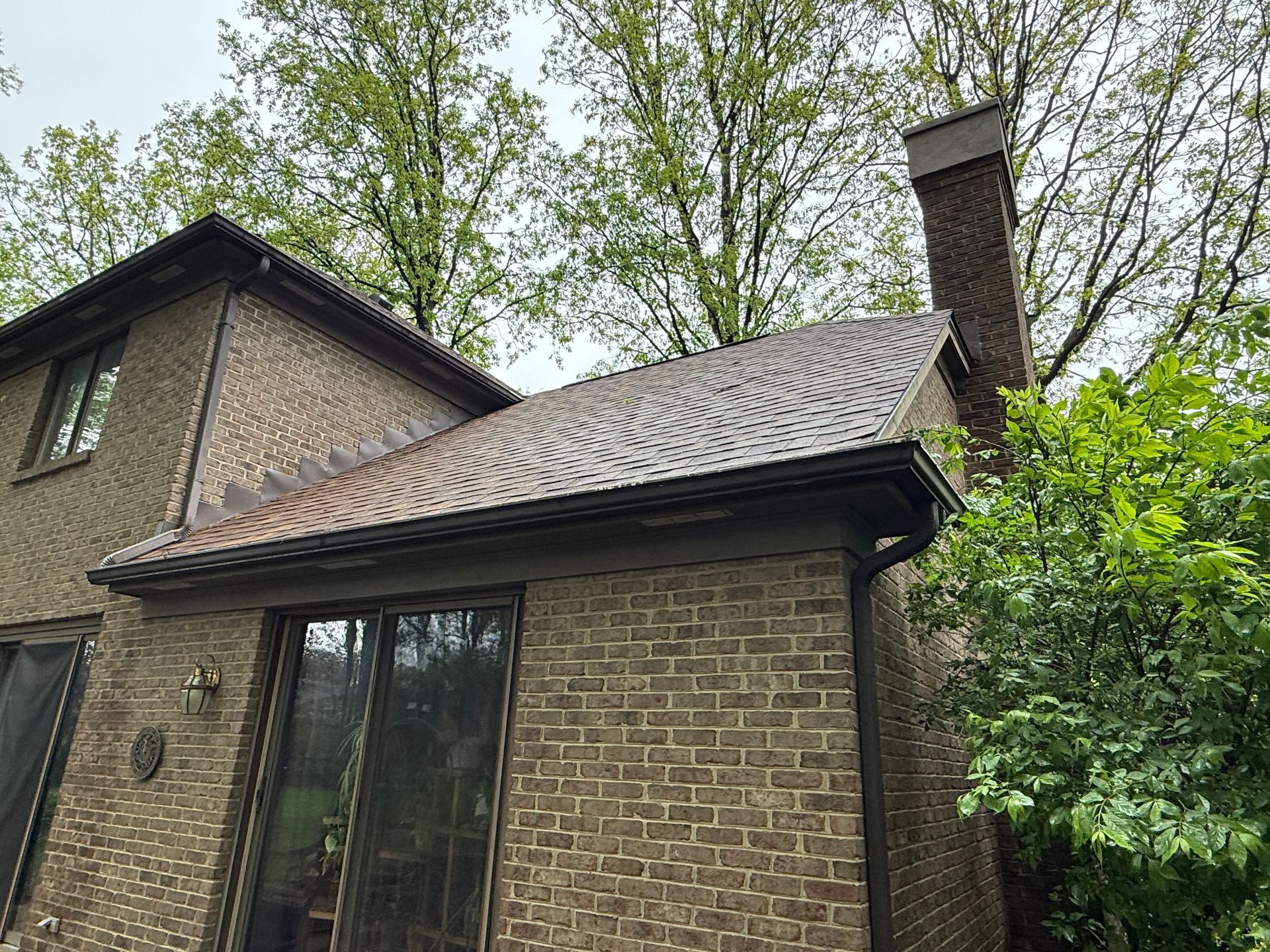 Brick house with curved shingle roof, chimney, and trees overhead