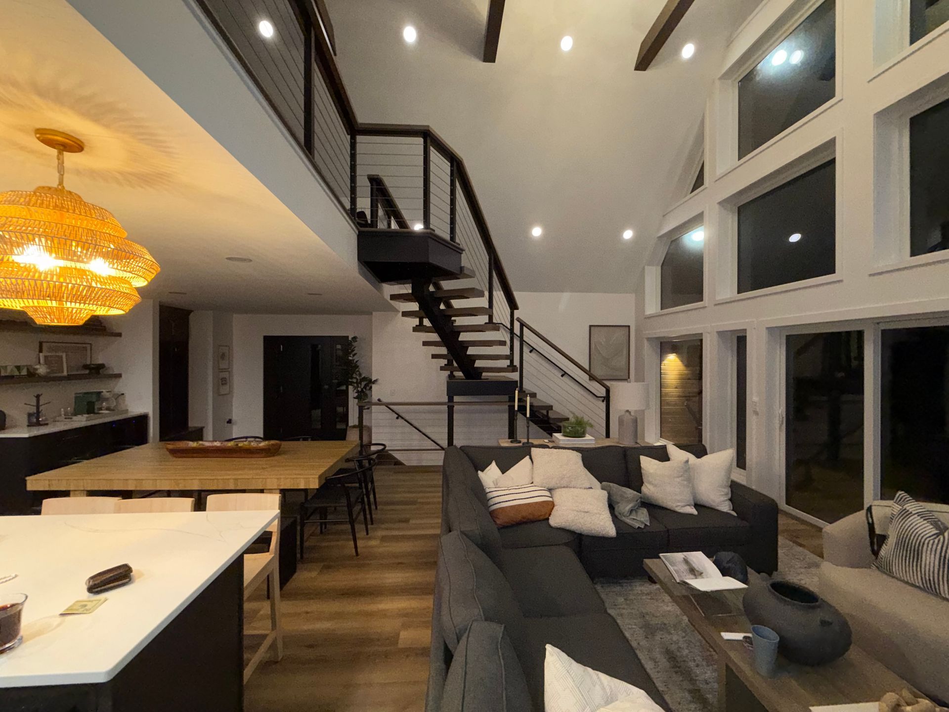 Modern open-plan living room with black staircase, large windows, gray sofa, and warm pendant lighting